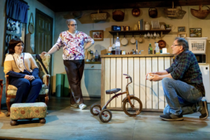Three actors look at each other in a living room. A tricycle is in the center.
