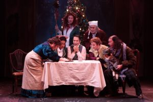 The cast leans in together around the Cratchit table with Scrooge and the Ghost of Christmas Present looking on.