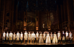 The company of Hamilton stands in a line across the front of the stage.