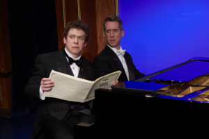 Two pianists sit at the piano, making an expression of stress.