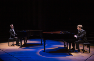 Two pianists sit facing on another at their instruments, bathed in blue light.
