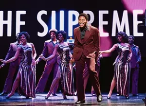 Eddie Kendricks sings midstage with his bandmates and the Supremes behind him. Temptations and Supremes are listed on the large light sign behind the performers.