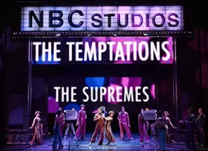 The Temptations sing and dance with the Supremes, surrounded by various video cameras. Temptations and Supremes are listed on the large light sign behind the performers.