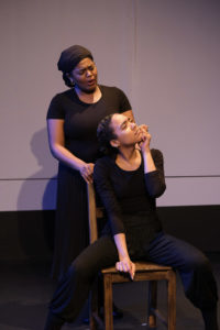 A woman stands behind a chair, pondering, while a dancer sits on it in front of her.