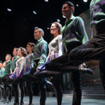 The Riverdance Dancers form a straight line across the front of a stage as they smile and sing.
